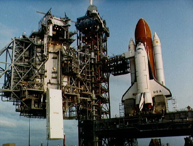 With the Space Shuttle waiting on launch pad 39A, the satellite toting payload canister is being hoisted to the environmentally clean payload…