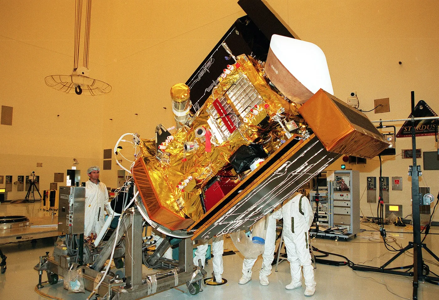 Workers in the Payload Hazardous Servicing Facility watch as the Stardust spacecraft is lowered before deploying panels for lighting tests.