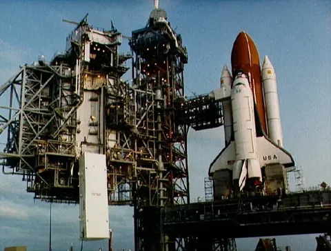 With the Space Shuttle waiting on launch pad 39A, the satellite toting payload canister is being hoisted to the environmentally clean payload…