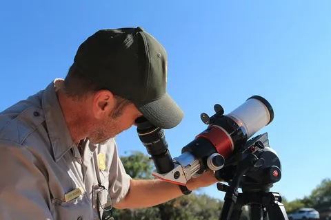 A park ranger wearing a grey shirt and green ball cap looks through a small telescope. A park ranger looks through a solar telescope viewfinder to…