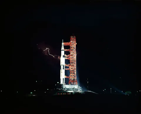 Apollo 15 at launch pad at night with lightning in the distance.
